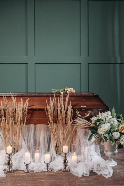 A wooden funeral casket decorated with a small bouquet of pink and white flowers resting on top, set against a green paneled background. In front of the casket, there are beige dried grasses arranged 