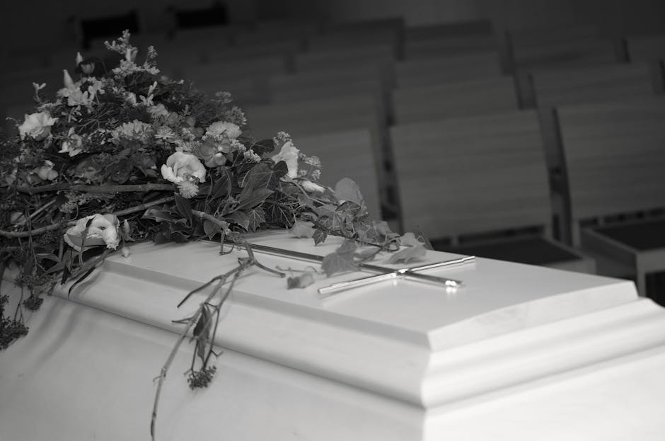 A closed white casket draped with a large floral arrangement featuring white roses, lilies, and greenery, placed on top of the casket. The bouquet includes a mix of fresh, textured petals and sturdy s