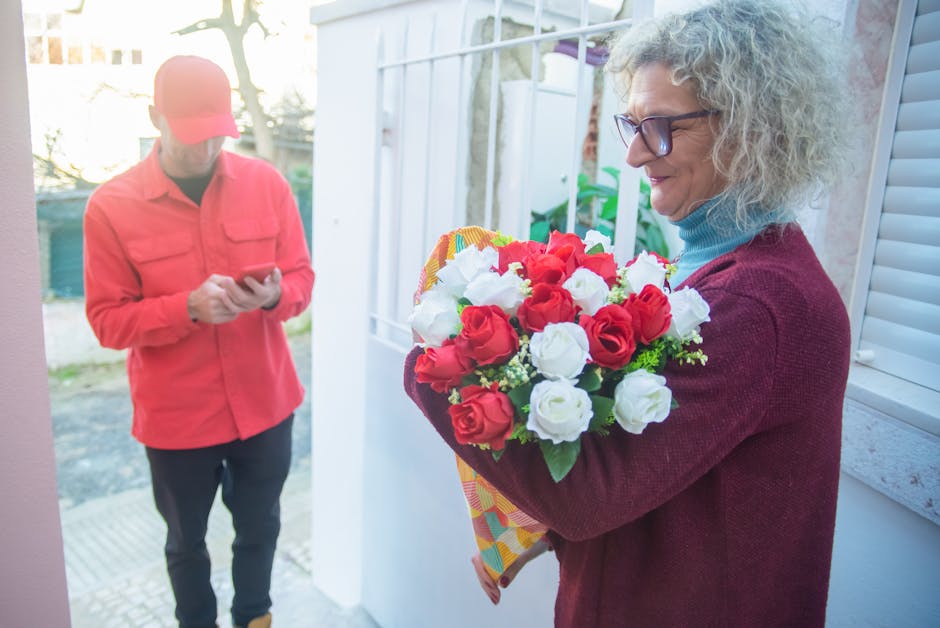 A woman with curly gray hair and glasses holds a vibrant bouquet of fresh roses, predominantly red and white, wrapped in bright patterned paper. She stands outdoors near a white door, with natural day