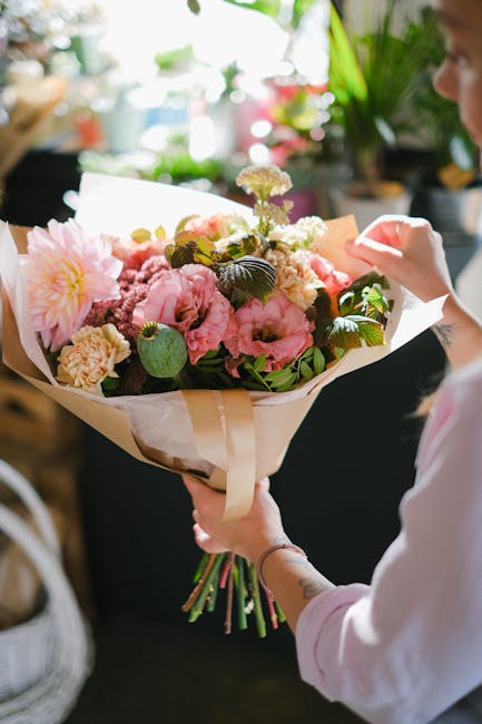 A person holding a large bouquet of fresh flowers, featuring pink peonies, white and light purple blooms, and dark green foliage, wrapped in light beige paper with a satin ribbon. The bouquet is being