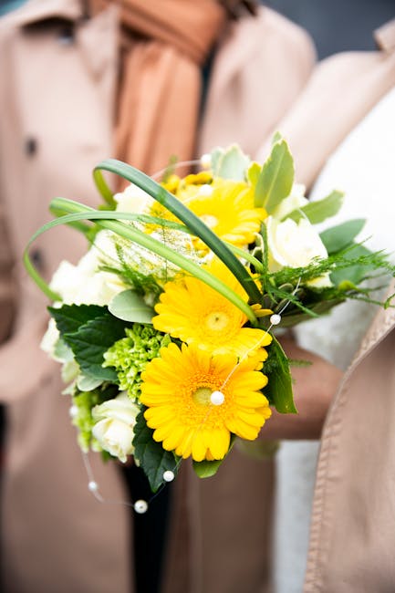 A floral arrangement featuring vibrant yellow gerbera daisies, white roses, and green foliage held by a person dressed in a beige coat and peach scarf. The bouquet is composed with lush leaves and del