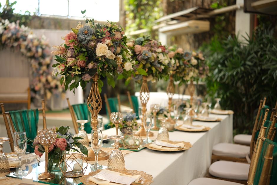 A long dining table decorated with elegant floral arrangements featuring pastel pink, cream, and soft blue flowers, including roses and hydrangeas, placed in tall, gold ornamental stands. The table se