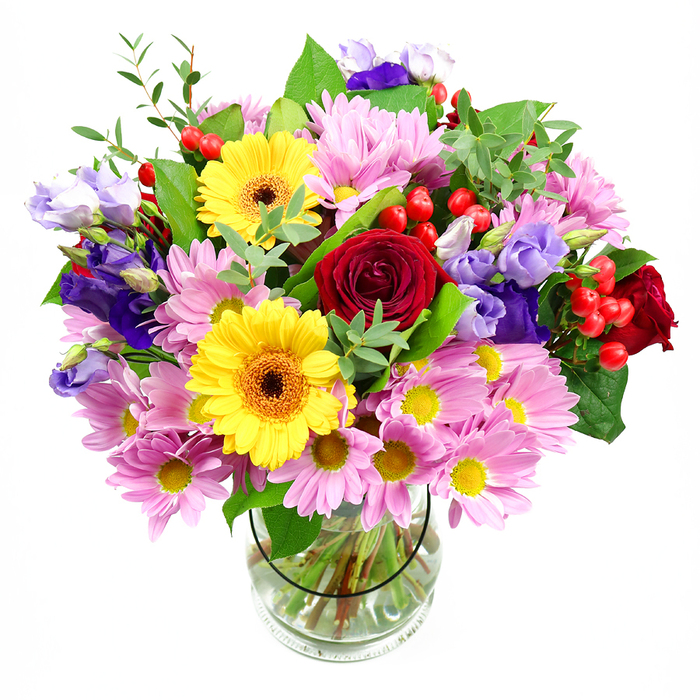 In this photograph, a vibrant mixed bouquet rises gracefully from a transparent glass vase, the whole arrangement sharply outlined against a clean white background. The florist has composed the design in a loose, rounded shape, with deep red roses forming the central anchor. These roses, with their plush, velvety petals spiralling inward, bring an immediate sense of romance and depth, almost like a quiet nod to timeless love stories shared in Crouch End cafés. Around them, generous clusters of pink daisies or chrysanthemums soften the look; each bloom is made up of many slender petals surrounding a neat yellow eye, creating a gentle halo of pastel colour that contrasts beautifully with the stronger reds. Two bright yellow gerbera daisies emerge confidently from the mix, their large, flat faces and rich brown-orange centres acting like cheerful bursts of sunshine, lifting the entire palette. Woven between these focal flowers are stems of lisianthus in hues of purple and lavender, their slender, bell-shaped blooms and softly ruffled edges introducing an elegant, vertical rhythm to the bouquet. Here and there, small clusters of glossy red hypericum berries appear like scattered jewels, adding dimensional interest and a modern textural note. A wealth of greenery surrounds everything, from broad, dark green leaves providing structure at the base to finer, light green foliage that extends outward, giving the arrangement a natural, airy movement. The clear vase reveals the carefully spiralled stems immersed in fresh water, showcasing professional skill and an eye for detail. The mood of the bouquet is bright, warm, and celebratory, perfect for placing on a dining table just off Crouch End Broadway, as a housewarming gift near Highgate, or as a same-day delivery from Crouch End Florist to someone who needs a burst of colour and joy in their everyday space.